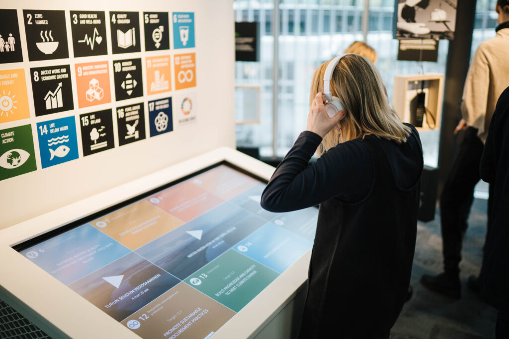 Henkilö kuulokkeet päässä tutustumassa näytöltä Turun kestävän kehityksen tavoitteisiin.