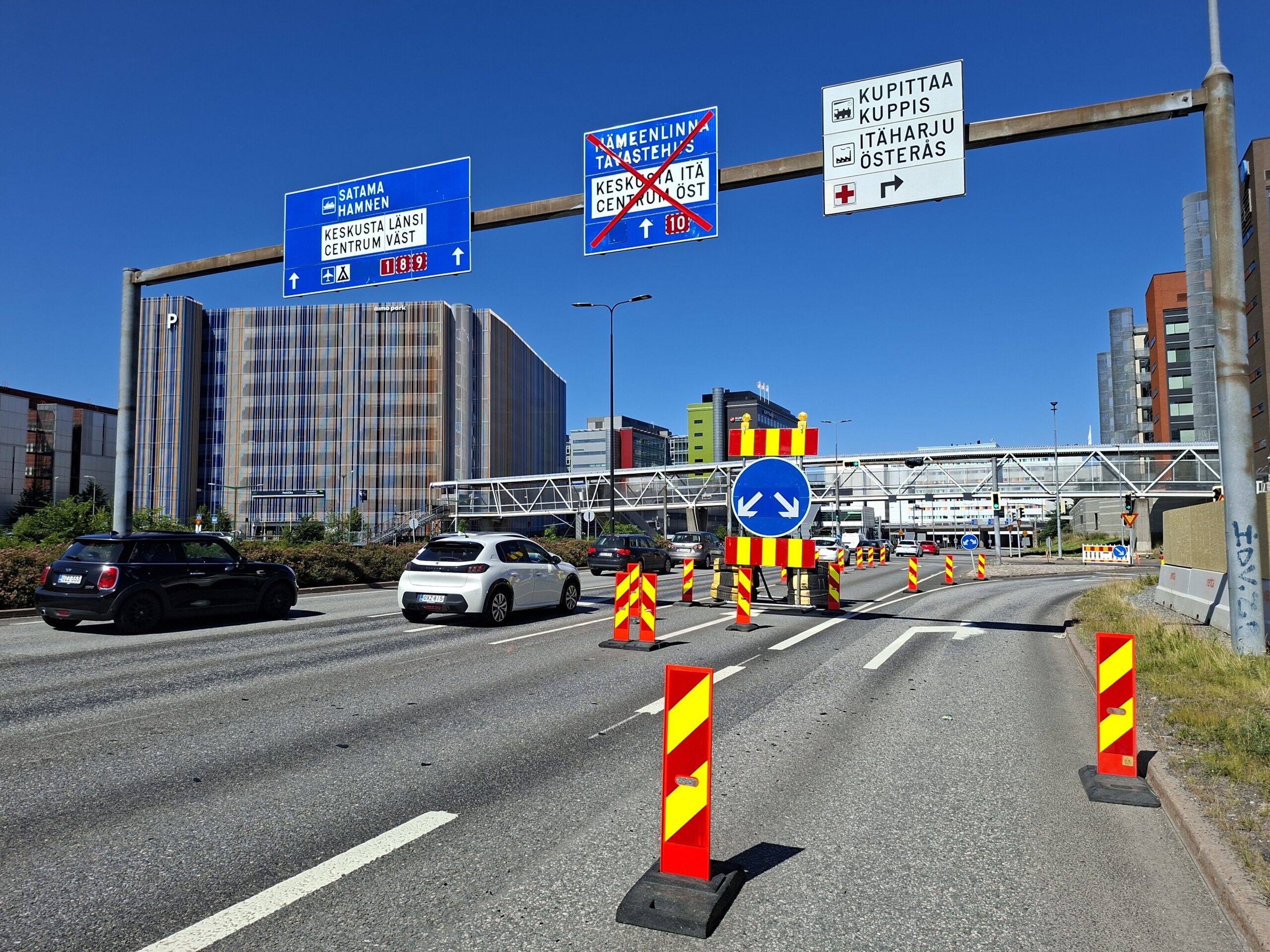 Kolmekaistaiselle tielle on työmaan sulkulevyillä kaistajärjestelyitä, yksi kaista on suljettu ja käytössä olevat kaistat osoitetaan merkillä. Tiellä siesoo autoja liikennevaloissa. Taustalla Turun Kupittaan rakennuksia ja siltoja.