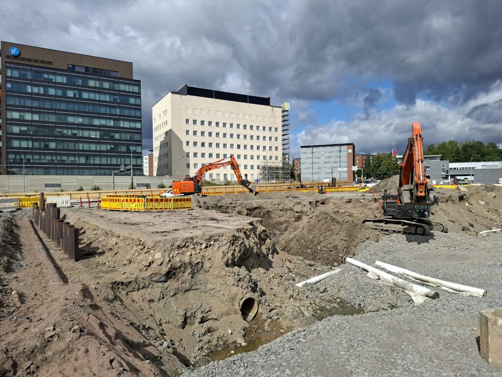 Työmaalla kaksi kaivuria, jotka kaivavat maata. Ympärillä useampia eri kaivantoja, vasemmalla kohoaa matala metallinen tukiseinä. Aluetta on aidattu. Taaempana kohoaa Kupittaan rakennuksia.